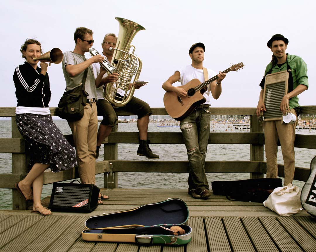Band, bestehend aus vier Männern und einer Frau, fröhlich gestimmt, auf einem Steg, mit ihren Instrumenten.