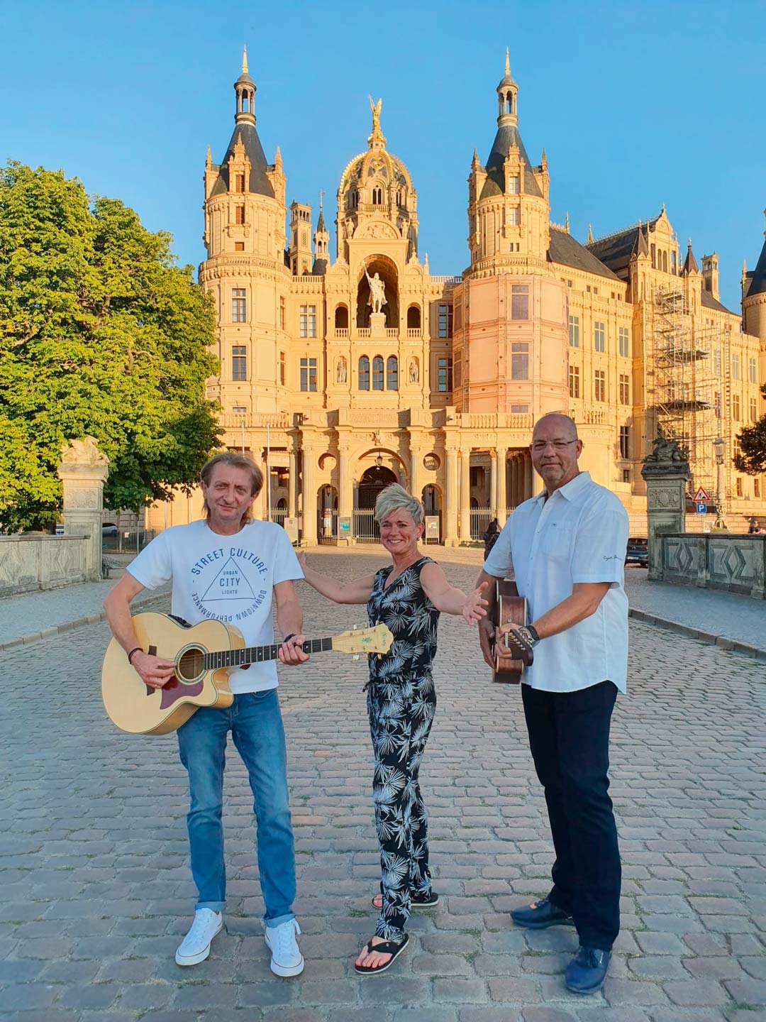 Ein Trio, bestehend aus zwei Männern und einer Frau, mit ihren Instrumenten vor dem Schweriner Schloss