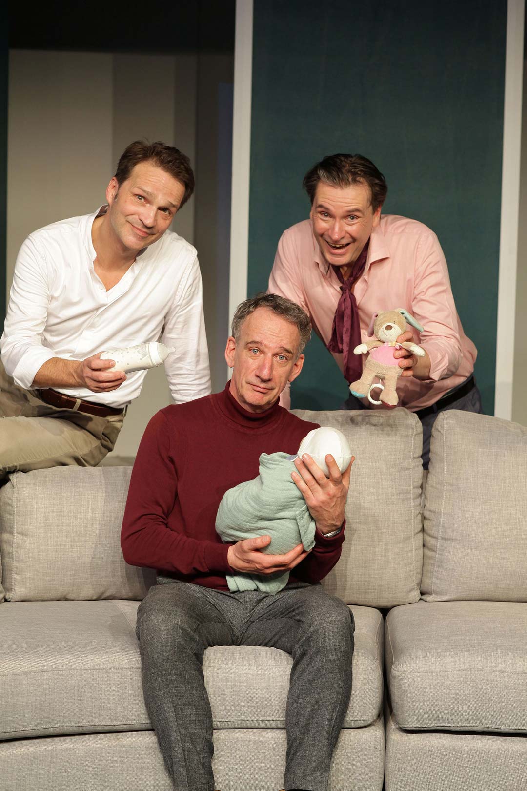 Drei Männer, davon einer auf der Couch sitzend, das Baby im Arm, während die anderen zwei sich leicht auf die Couch lehnen