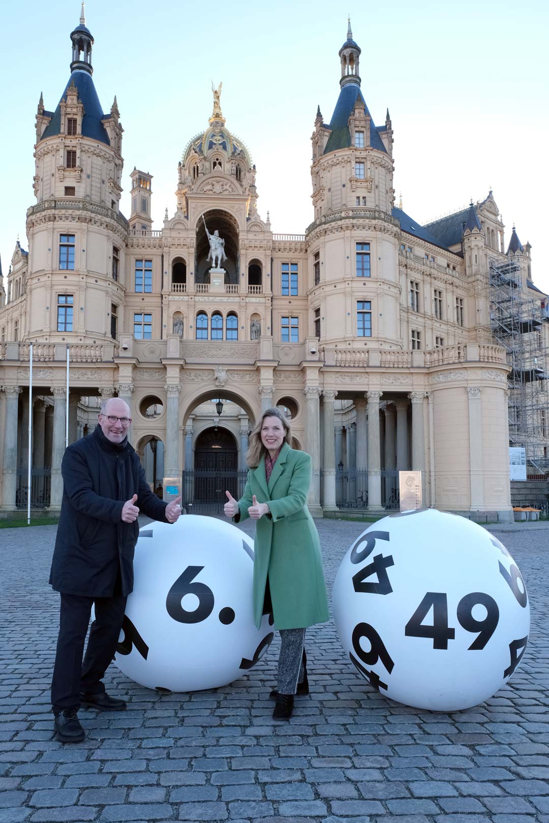 Zu sehen sind eine Frau und ein Mann vor dem Schweriner Schloss mit zwei großen Lottozahlen